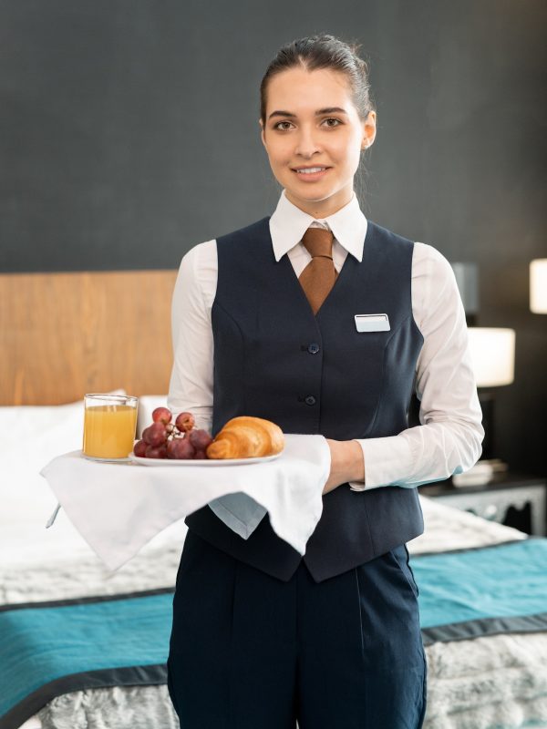 happy-young-elegant-female-worker-of-hotel-holding-tray-with-breakfast-against-bed-in-hotel-room.jpg